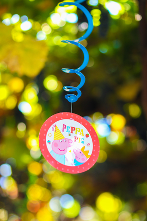 Fragment of decoration, decoration thematic of childrens holiday, birthday, photo colorful round medals with image of Peppa pig, suspended on blue spiral and decorated with yellow ribbons, against background of green foliage in courtyard outdoors. Conceptの写真素材