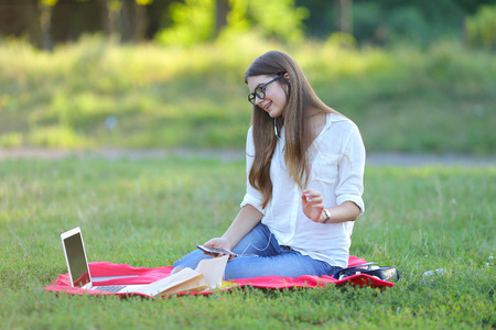 girl listening to music on the nature of the glasses. in the park for a laptop unravels headphones and holding a mobile phoneの写真素材