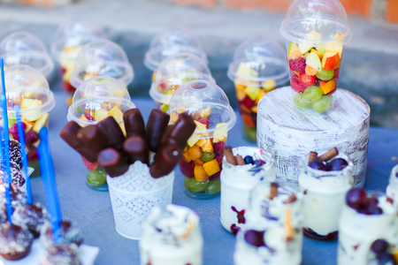 In photo depicts variety of goodies, delicious food, clear jars with cottage cheese soufflÃÂ© with layer of pastry topped with fruit and straws, chocolate long candy skewers in patterned white small vase, chocolate balls in coconut flakes with blue strawの写真素材