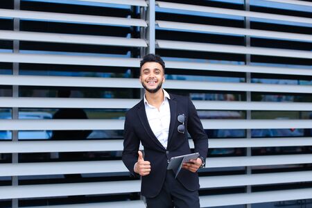 Young handsome businessman Arab Muslims is in the business center in black elegant dress on building background. Businessman man male looking waiting concept and smiling.の写真素材