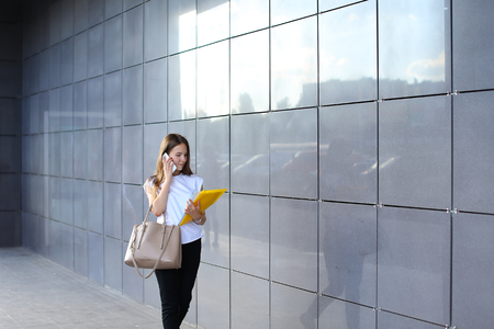Successful young business woman walks and talks on phone, holds and looks at documents in yellow folder, smiling on background of gray wall of shopping business center. Girl dressed in white shirt, black pants, beige shoes and keeps on hand beige bag. Conの写真素材