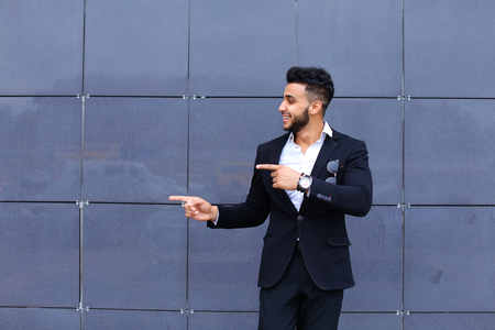 Young handsome businessman Arab Muslims is in the business center in black elegant dress on building background. Businessman man male looking at time walking goes and smiling.の写真素材