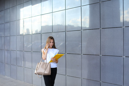 Serious female girl businesswoman talks on phone, holds and looks at documents in yellow folder on background of gray wall of shopping business center. Girl dressed in white shirt, black pants and keeps on hand beige bag. Concept of business meeting, busiの写真素材