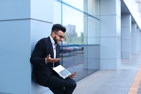 Entrepreneur uses  laptop wearing sunglasses. Young handsome businessman arab muslims in business center wearing dressed in black elegant suit on building background.の写真素材