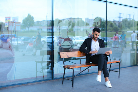 Entrepreneur uses laptop wearing sunglasses. Young handsome businessman arab muslims in business center wearing dressed in black elegant suit on building background.の写真素材