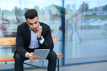 Entrepreneur smiling man  puts on sunglasses uses tablet and looks at. Young handsome businessman arab muslims in business center wearing dressed in black elegant suit on building background.の写真素材