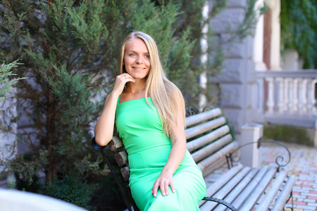 Beautiful female girl posing for photo, holding hand near face and smiling, looking away and sitting on gray wooden bench on background of coniferous tree in  courtyard outdoors. Girl with blond long hair dressed in green sundress below knee. Concept of aの写真素材
