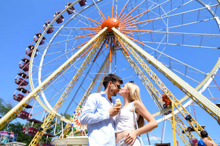 Cute young people, newlyweds kissing, hugging and smiling, looking into eyes of each other and posing for photos, guy holding ice cream on background of large and colorful ferris wheel at an amusement park outdoors. Girl with long blond hair dressed in brの写真素材