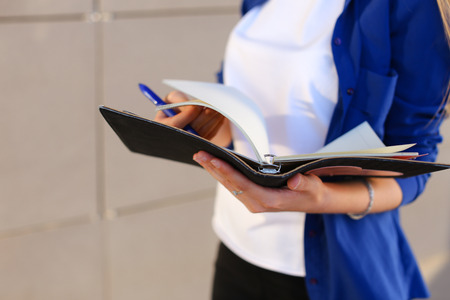 Girl holds in hands diary, black notebook with sheets and blue pen and leafs through pages. Young woman dressed in white T-shirt and bright blue shirt with pocket on arm bracelet.Concept of organization and planning of day, notebook, homework, busy dealerの写真素材