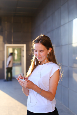 Business attractive girl young business woman holds white telephone, listens to music, uses phone to work, solves problems, smiles and stands in profile on background of gray wall of business center. Girl with long light brown hair dressed in white shirt の写真素材