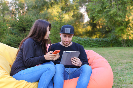 Pair of lovers of young attractive people sitting in soft yellow and red chair, man uses gadget that keeps in arms, female holds plastic card, coupon. Young people cute communicate, plan to travel, choosing interior of apartment, she shows on flyer and guの写真素材