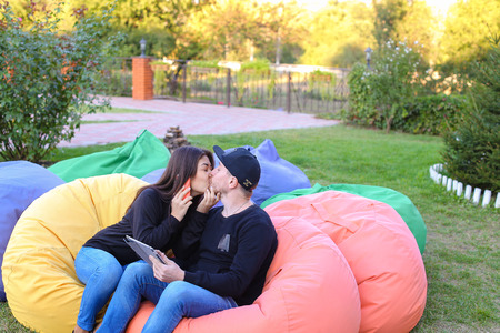 Pair of lovers of young attractive people sitting in soft yellow and red chair, man uses gadget that keeps in arms, female holds plastic card, coupon. Young people cute communicate, plan to travel, choosing interior of apartment, she shows on flyer and guの写真素材