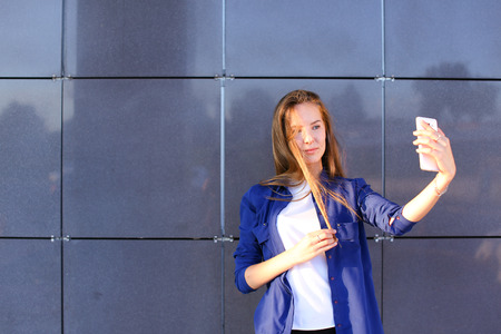 Charming cute lady girl student photographed, makes selfie photos and smiling at camera on background of business center on outdoors. Girl with long light brown hair dressed in white T-shirt and blue shirt. Concept of Concept of takes selfie, cellphone, mの写真素材