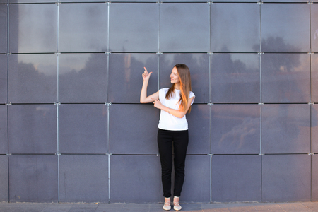 Beautiful young energetic female girl raised arms and points to advertising space, worth exactly in full growth and smiles on background of gray wall of modern business center.Girl dressed in white shirt, black pants, beige shoes. Concept of pointer to, aの写真素材