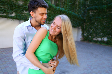 Enamoured guy and girl, engaged couple looking into each others eyes, smiling and embracing, posing for photos and stand in patio of house on background of overgrown with vines and leaves outdoors. Girl with long blond hair dressed in green sundressguy drの写真素材