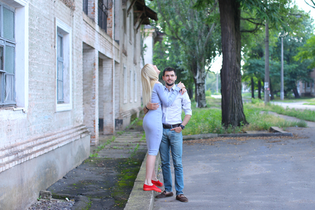 Girlfriend and boyfriend standing next to each other, posing, talking and laughing, hugging on street. Girl dressed in midi gray dress and red slip-on, guy straight, keeping hands in pants pockets and looking directly into camera, he dressed in blue jeansの写真素材