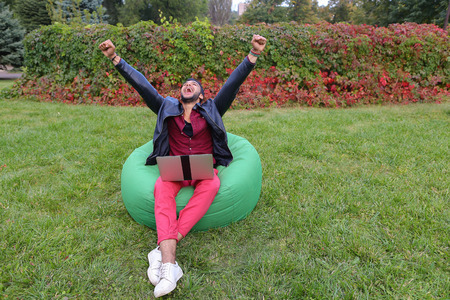 Perspective Young Arabic Guy, Businessman, Student Sits With Laptop in Soft Green Chair, Looks at Computer Screen and Smiling, Rejoice Success, Winning on Stock Exchange, Working Online on Background of Green Grass in Garden Outdoors. Bearded Man Dressed の写真素材