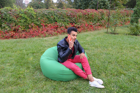 Business Muslim Arabian Guy Businessman, Student Gets From Jacket Pocket Cellphone and Answers Call, Talking on Mobile, Solves Business Issues, Talking With Friend, Smiling and Laughing, Removes Sunglasses and Sitting in Comfortable Green Chair in Bloominの写真素材