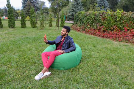 Young Handsome Man Arab Businessman, Student Holding Mobile Phone, Gadget and Calls, Communicates on Skype For Video Calling, Smiling and Sitting in Comfortable Green Chair in Garden Outdoors. Bearded Man With Dark Hair Dressed in Black T-Shirt, on Top Maの写真素材