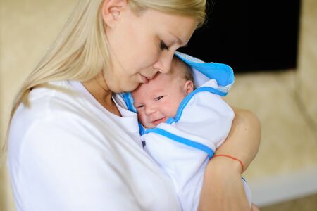 Young female lulls and sings songs for child, gently hugging and talking with kid, baby cries and looks at mother, located in mothers arms in bright room. Woman blonde European appearance dressed in white T-shirt, newborn baby wrapped in white blanket witの写真素材