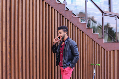 Confident Attractive Young Guy Muslim Businessman Goes Slowly Along Wall, Holding Phone in Hand and Talks, Resolve Issues, Discuss Future Plans and Smiling, Holds Hand in Pocket, Stands on Background of Wooden Panel Stairs Near House Outdoors. Bearded Manの写真素材