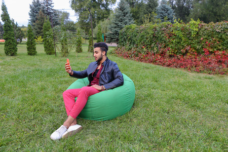 Young Handsome Man Arab Businessman, Student Holding Mobile Phone, Gadget and Calls, Communicates For Video Calling, Smiling and Sitting in Comfortable Green Chair in Garden Outdoors. Bearded Man With Dark Hair Dressed in Black T-Shirt, on Top Maroon Shirの写真素材