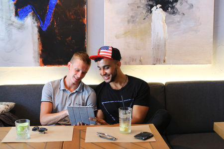 Two male friends sitting in cafes guy using phone technology socialize, discuss, watch, share secrets, waiting for the order, socialize, smiling, satisfiedの写真素材