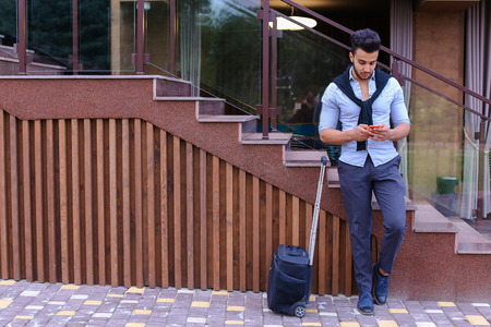 Young man businessman Arabian beautiful appearance is black travel bag on small wheels and is waiting for taxi, on journey or business trip, using phone and check their seats in airplane online, reserve hotel, one leg held up other, and based on stairs neの写真素材