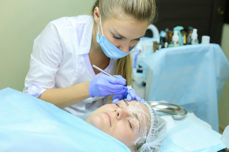 Young blonde woman professional, engaged in reconstruction work responsibly, eyebrows manual method performs procedure, using thinnest needle and maniples, draws most natural eyebrow, side by side on couch and ready for client procedure in cosmetic white の写真素材