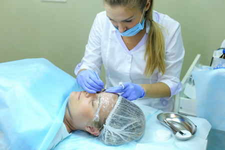 Master of mikrobleyding young attractive girl evaluates correct execution of work, check with help of special device density of drawing and color of hairs of eyebrows, brings to end of last stage in bio tattooing eyebrows. Master dressed in white robe, blの写真素材