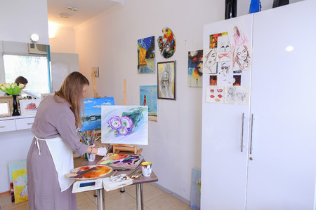 Female Artist Paints Picture Uses Brush and Oil Paints on Canvas. Woman Draws Purple Flowers on White Background, stands with back to camera and Answers to Important Phone Call, Continually Talks on Smartphone. Woman With Long Light Brown Hair Dressed Shtの写真素材