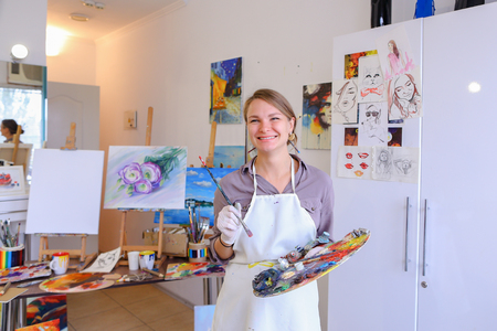 Cute female painter posing, laughing and fooling around in front of camera and smiling with brushes and colorful palette in hand, standing in studio with white art paintings on walls. Girl European appearance advertises art school for adults and children,の写真素材