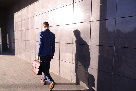 Handsome young male guy entrepreneurstudent hurry on business meeting, returns from lunch, going home after work, looks at his watch put on by on  hand, late and goes on background of gray wall of business center outdoors. Stylish man dressed in blue busiの写真素材