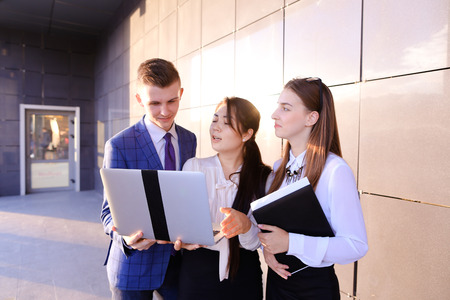 Modern young man and two attractive smart young women, business people, students communicate, holding laptop and study work plan, familiarize with presentation, teach each other new programs, solve important problems and workloads holding folder with docuの写真素材