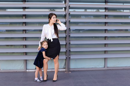 Elegant young female woman, young modern mom, business woman holding mobile phone hand and talking on cellphone, solves problems, negotiates business meeting with partners, spends time girl daughter, talking, smiling at her, little girl hugging her motherの写真素材