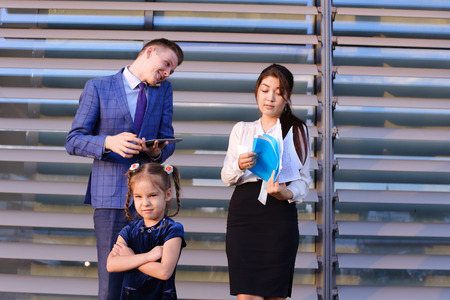In  foreground stands and frowns, angry with arms crossed in front of little girl, child,  in background successful young handsome mans man, young father and modern woman, young mother hold gadgets in hand, mobile phones and talking to them, solve operatiの写真素材