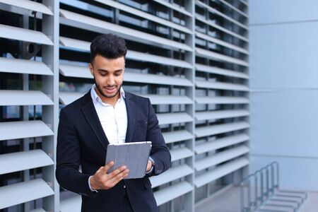 Young handsome businessman Arab Muslims is in the business center in black elegant dress on building background. Businessman man male looking waiting concept and smiling.の写真素材