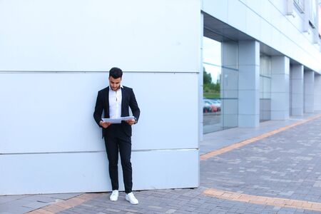 Young tall handsome arab entrepreneur guy student holds and studies, reads paper or documents, stands in full growth directly into camera near gray wall of business center. Bearded man dressed in classic suit, black pants, jacket , white shirt and sneakerの写真素材