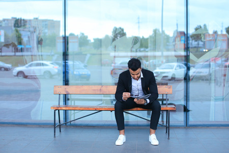 Entrepreneur smiling man  puts on sunglasses uses tablet and looks at. Young handsome businessman arab muslims in business center wearing dressed in black elegant suit on building background.の写真素材