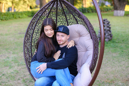 Couple in love swinging on chair in street among  large green park, discussing plans for future, share secrets and jokes, young people hugging each other gently. Guy dressed in black sweater, blue jeans, sneakers and black cap, girl with long black hair dの写真素材