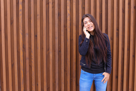 Cute girl with long hair cute talking on phone with friend or loved one, make an appointment. Women dressed in black sweater, blue jeans, black boots on platform and top dressed in jacket standing on background of brown wood panel walls near restaurant ouの写真素材