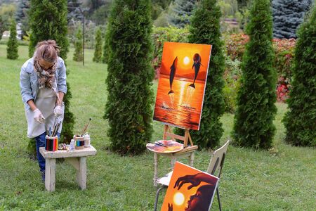 Artist cleans workplace, evaluate work that stands on easel, smiling, posing and looking in camera, standing in full length near paintings in park outdoors on background of coniferous plants in afternoon. Girl dressed in red sweater, dark jeans, white aprの写真素材