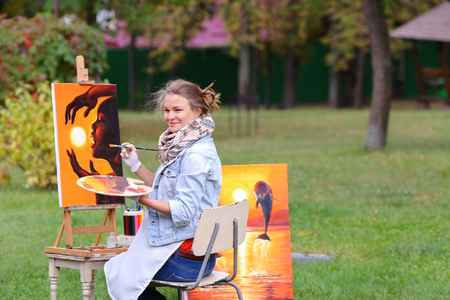 Young cute woman artist  finishes painting which depicts woman who keeps  sun on orange background, sitting with his back to  photographer and  behind wooden easel, holds palette smeared in paint in large park green day outdoors.Girl dressed in red sweateの写真素材