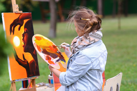 Young cute woman artist  finishes painting which depicts woman who keeps  sun on orange background, sitting with his back to  photographer and  behind wooden easel, holds palette smeared in paint in large park green day outdoors.Girl dressed in red sweateの写真素材