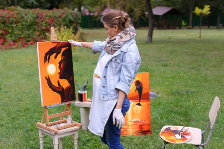 Female European appearance artist evaluates picture and removes from easel, holds and posing with picture which depicts woman with sun in hands made orange smiling and standing in full growth on background of lawn and angered by conifers in large park in の写真素材