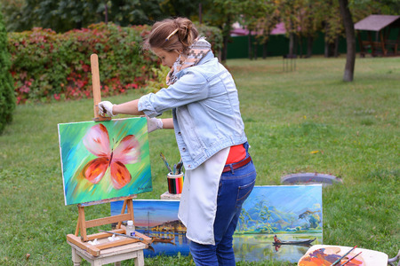 Female European appearance artist evaluates picture and removes from easel, holds and posing with picture which depicts colorful butterfly, smiling and standing in full growth on background of lawn and angered by conifers in large park in open air. Girl dの写真素材