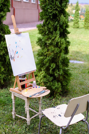 Photo depicts wooden easel on which stands white canvas with painting started, Wooden palette with paints and bank decorated with colored pencils which stands with brush located in park outdoors. Concept of art materials, oil or acrylic paint, professionaの写真素材