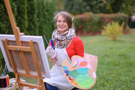 Cute female painter posing, laughing and in front of camera , draws and smiling with brushes and colorful palette in hand, sits on chair near easel in green park outdoor.  Girl with brown hair dressed in red jamper, dark denim overalls and white apron, chの写真素材
