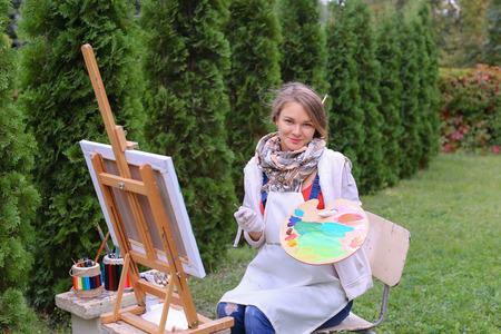 Cute female painter posing, laughing and in front of camera , draws and smiling with brushes and colorful palette in hand, sits on chair near easel in green park outdoor.  Girl with brown hair collected in beam dressed in light jacket, red jacket, dark deの写真素材