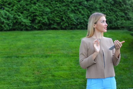 Stylish young girl of european appearance views all around and smiles little smile, thinking about something pleasant. Girl dressed in light blouse, beige jacket and blue pants, stands on background of green lawn and coniferous plants outdoors. Concept ofの写真素材